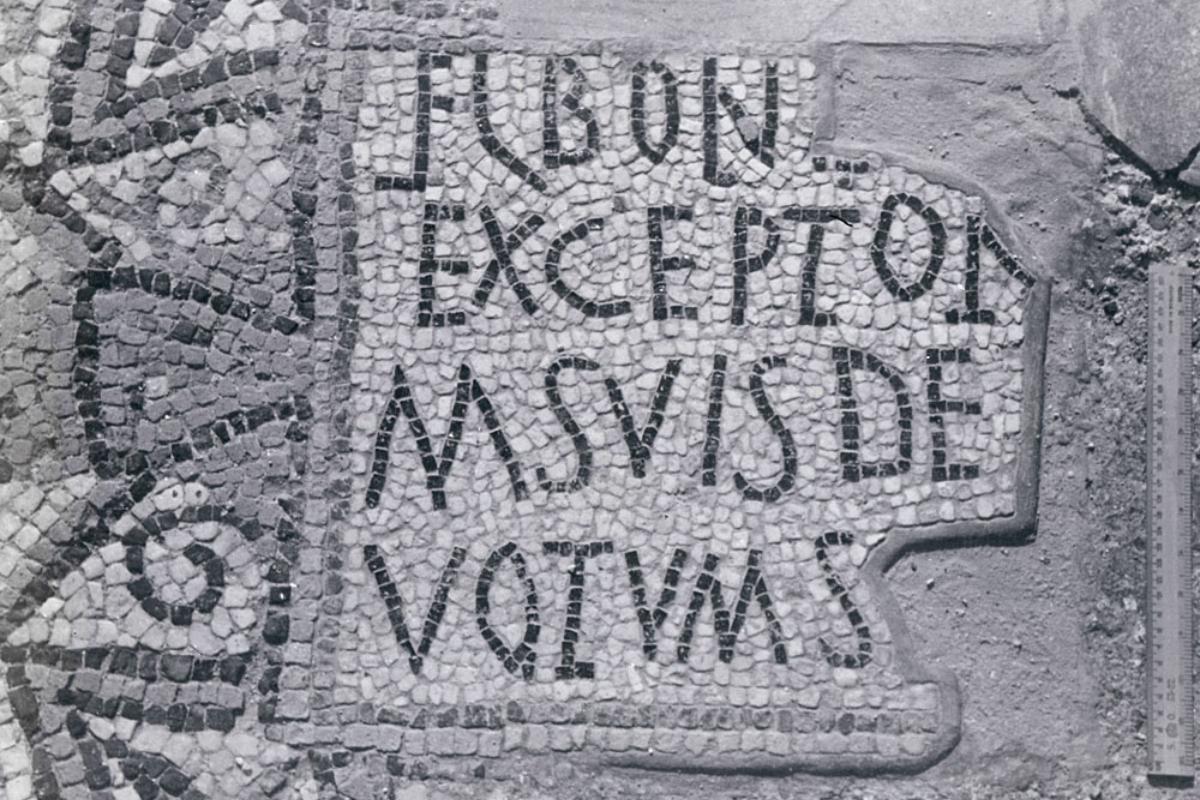 A black and white photograph of an ancient mosaic inscription with Latin text, surrounded by geometric patterns on a stone floor.